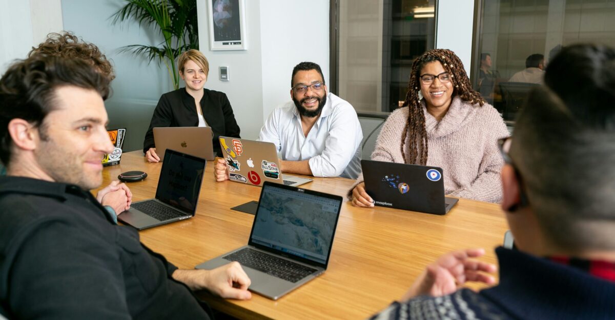 four people all on laptops two men and two women listen to person talking in a board meeting