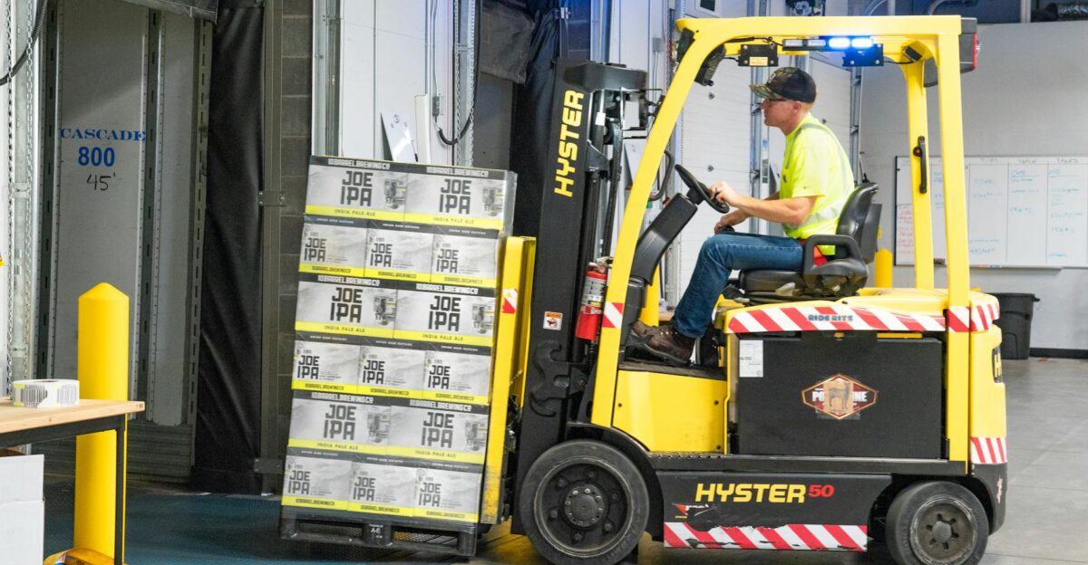 A worker drives a Hyster forklift moving Joe IPA boxes in a warehouse