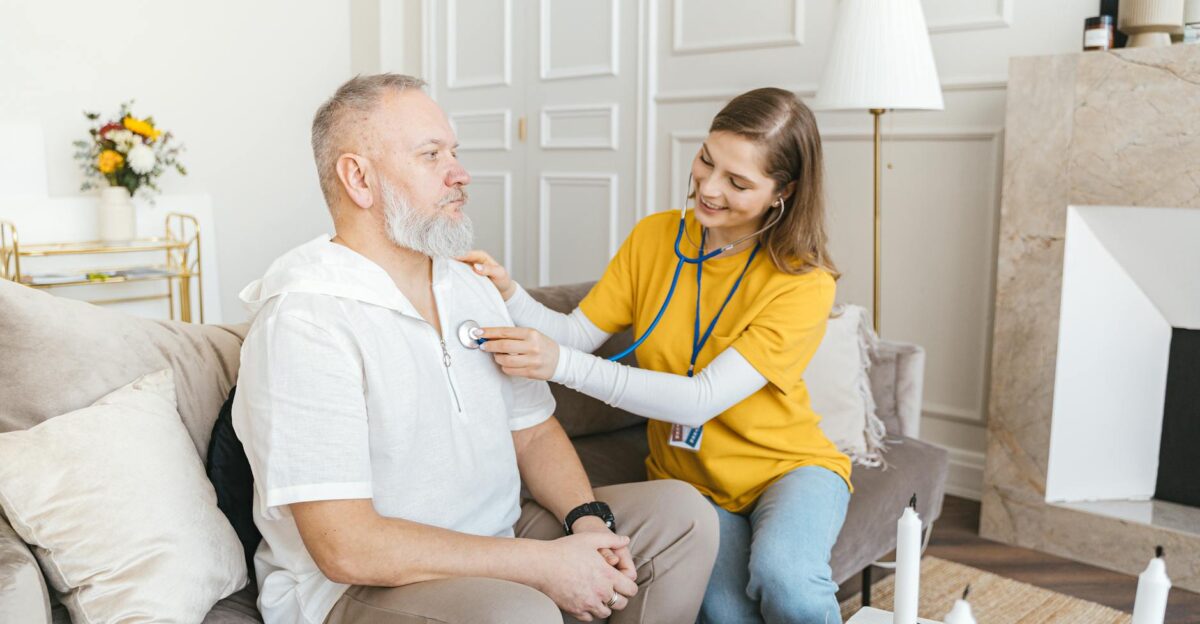A nurse uses a stethoscope for a home check-up on a senior adult in a cozy living room